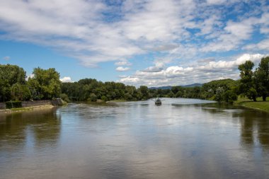 Vah nehrinin sakin suları, güneşli bir yaz gününde mavi gökyüzünü beyaz bulutlarla yansıtıyor. Sahilde taze yeşillik. Piestany, Slovakya.