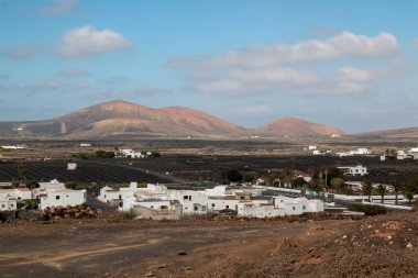 Şarap üzümlerinin ekildiği karanlık volkanik bir toprak manzarası. Geleneksel beyaz binalar. Arka plandaki dağ. Kışın beyaz bulutlu mavi gökyüzü. La Florida, Lanzarote, İspanya.