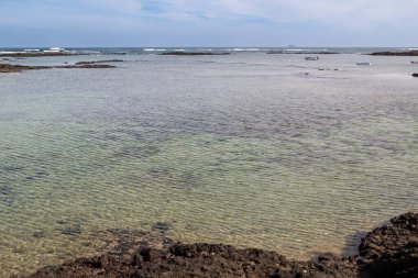 Beyaz kumlu ve sığ sulu bir sahil. Siyah volkanik küçük kayalar. Kışın hafif bulutlu mavi gökyüzü. Caleton Blanco, Lanzarote, Kanarya Adaları, İspanya.