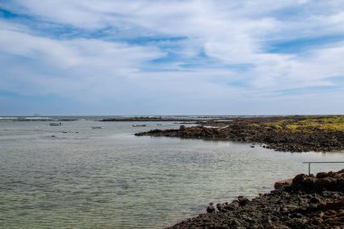 Beyaz kumlu ve sığ sulu bir sahil. Siyah volkanik küçük kayalar. Kışın hafif bulutlu mavi gökyüzü. Caleton Blanco, Lanzarote, Kanarya Adaları, İspanya.