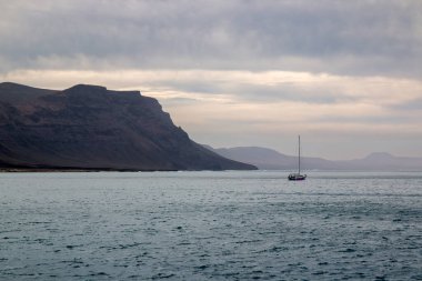 Adanın kuzeyindeki kayalıklardan ve volkanik kayalıklardan bir tekne görüntüsü. Küçük tekne. Kışın beyaz bulutlu mavi gökyüzü. Mirador del Rio, Lanzarote, Kanarya Adaları, İspanya.