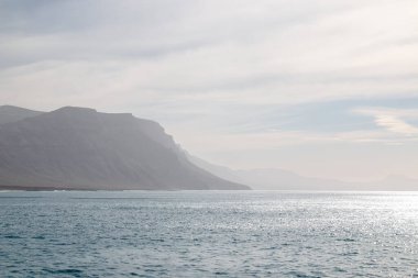 Lanzarote 'un kuzeyindeki kayalıklar sisli. Atlantik okyanusunun sakin suları. Kışın bulutlu bir gökyüzü. Caleta del Sebo, La Graciosa, Kanarya Adaları, İspanya.