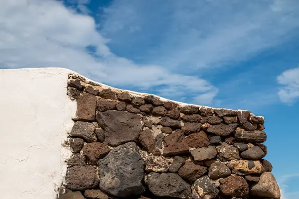 stock image Wall made of various brown tones of volcanic stones. Another part with a white facade. Blue sky with white clouds. Caleta del Sebo, La Graciosa, Canary Islands, Spain.