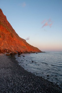 Altın saat volkanik kayayı parlak turuncu bir renge boyuyor. Atlantik okyanusunun sakin suları. Mavi gökyüzü ve biraz ışık bulutu. Paul do Mar, Madeira, Portekiz.