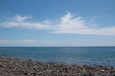 Sahildeki volkanik çakıl taşlarının koyu rengi. Atlantik okyanusunun mavi sakin suyu. Parlak mavi gökyüzü ve ışık yapılandırılmış bulutlar. Madalena do Mar, Madeira, Portekiz.