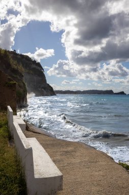 Uçurumlar çeşitli bitkilerle kaplı. Yaz sonlarında dalgalı iyonik bir deniz manzarası. Panoramanın tadını çıkarmak için beton boş banklar. Beyaz bulutlu mavi gökyüzü. Yialos Sahili, Kuzey Korfu, Yunanistan