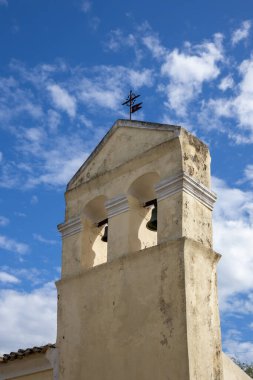 Geleneksel bir ortodoks kilisesinin çanı. Yolun yanındaki küçük bina. Beyaz bulutlu mavi gökyüzü. Acharavi, Korfu, Yunanistan.