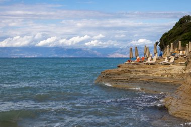 Kum taşı kayalıkları, taze yeşilliklerle kaplı, bir körfez kanalı d Amour (Aşk Kanalı) oluşturur. Boş sandalyeler ve kapalı güneş şemsiyeleri. Beyaz bulutlu mavi gökyüzü. Sidari, Korfu, Yunanistan.