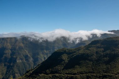 Dağlar ve vadiler çeşitli taze yeşilliklerle kaplıdır. Kayaların tepeleri bulutlarla ya da sisle kaplıydı. Yukarıdaki parlak mavi gökyüzü. Orta Madeira, Portekiz.