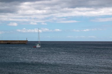 Limanda küçük bir yelkenli, Atlantik Okyanusu 'nun limanında yelkensiz yüzüyor. Arka planda iskele ve okyanusun ufku. Beyaz bulutlu mavi gökyüzü. Kanik, Madeira, Portekiz.
