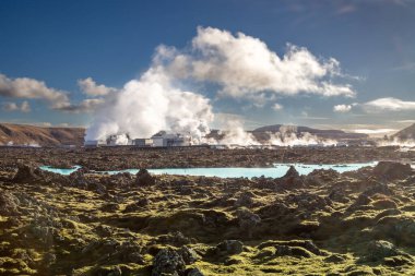 Yeşil yosunlarla kaplı lav tarlası. Bir gölün parlak mavi suyu, Blue Lagoon 'un bir parçası. Arka planda dağlar. Jeotermal buhar. Mavi gökyüzü. Grindavik, İzlanda.