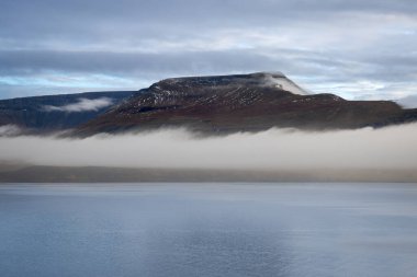 Calm water in a fjord. Mountains with a fog in the background. Cloudy sky during sunset. Akrafjall, Iceland. clipart