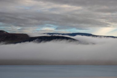 Calm water in a fjord. Mountains with a fog in the background. Cloudy sky during sunset. Akrafjall, Iceland. clipart