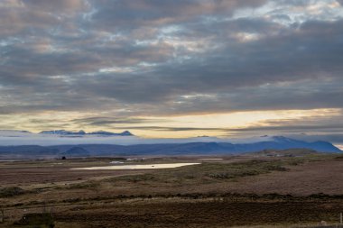 Sonbahar doğası. Zemin çimen, yosun ve yosunla kaplı. Gölün suyu. Arka planda sis olan dağlar. Gün batımı bulutlu gökyüzü. Avcılık / Hvalfjordur, İzlanda.