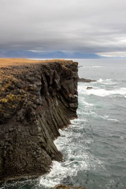 Atlantik Okyanusu kıyısındaki kayalıklar. Ufuktaki dağlar, kısmen sonbahar bulutlarıyla kaplı. Arnarstapi, Peninsula Snaefellsnes, İzlanda.