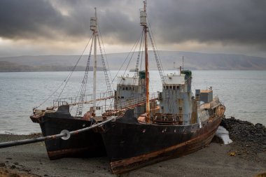 Fiyort kıyısında karaya vurmuş iki eski balina avcısı. Arka planda dağlar. Yoğun bulutlu gökyüzü. Hvalfjordur, Batı İzlanda.