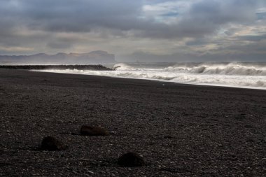 Atlantik Okyanusu 'nun kara volkanik kumsaldaki büyük dalgaları nefes kesici bir kontrastlık yaratıyor. Arka plandaki dağ. Bulutlu gökyüzü. Vikurfjara, Vik, İzlanda.