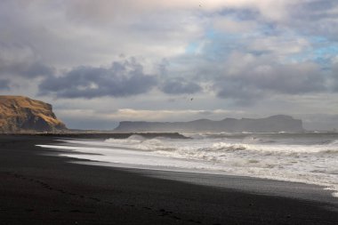 Atlantik Okyanusu 'nun kara volkanik kumsaldaki büyük dalgaları nefes kesici bir kontrastlık yaratıyor. Arka plandaki dağ. Bulutlu gökyüzü. Vikurfjara, Vik, İzlanda.