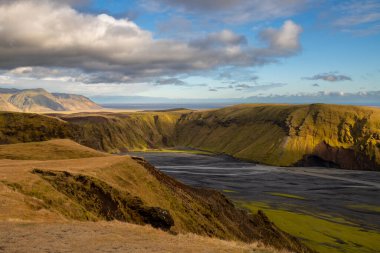 Nehrin dibindeki karanlık volkanik kum, sonbaharda neredeyse boş, sonbahar renklerinde yosun, liken ve otlarla kaplı dağlarla çevrili. Beyaz bulutlu mavi gökyüzü. Katla geopark, İzlanda.