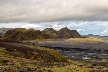 Nehrin dibindeki karanlık volkanik kum, sonbaharda neredeyse boş, sonbahar renklerinde yosun, liken ve otlarla kaplı dağlarla çevrili. Beyaz bulutlu mavi gökyüzü. Katla geopark, İzlanda.