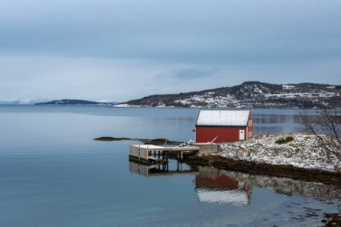 Sonbahar sonu ülkesi, karla kaplı. Norveç Denizi 'nin (Arktik Okyanusu) suyu, bulutlu mavi gökyüzünü yansıtır. Sahildeki kırmızı ev. Harstad, Norveç.