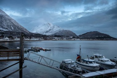 Norveç denizinin kıyısında küçük bir liman var. Ahşap iskele. Arka planda dağlar. Karla kaplı topraklar. Sonbaharın sonunda bulutlu bir gökyüzü. Kongsvika, Norveç.