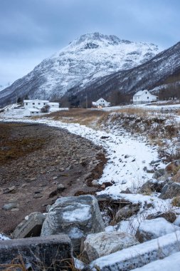 Norveç Denizi kıyısında. Sahildeki taşlar ve kayalar. Arka plandaki dağ. Birkaç ev. Karla kaplı topraklar. Sonbaharın sonunda bulutlu bir gökyüzü. Kongsvika, Norveç.