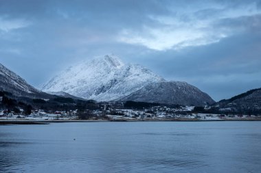 Norveç denizinin kıyıları kışın kısa bir günün mavi tonlarıyla arka planda dağlar. Karla kaplı topraklar. Sonbaharın sonunda bulutlu bir gökyüzü. Kongsvika, Norveç.