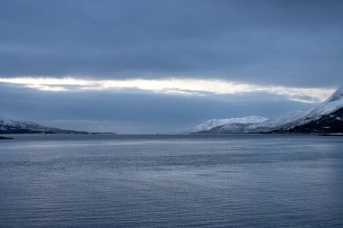 Norveç denizinin kıyıları kışın kısa bir günün mavi tonlarıyla arka planda dağlar. Karla kaplı topraklar. Sonbaharın sonunda bulutlu bir gökyüzü. Kongsvika, Norveç.