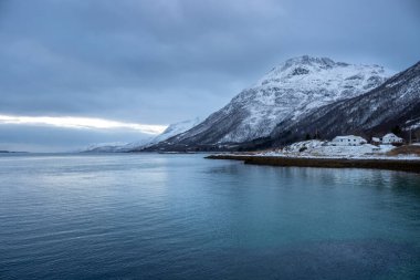 Norveç denizinin kıyıları kışın kısa bir günün mavi tonlarıyla arka planda dağlar. Karla kaplı topraklar. Sonbaharın sonunda bulutlu bir gökyüzü. Kongsvika, Norveç.