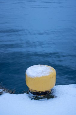 Fresh snow during a snowfall day. Yellow mooring bollard for boats on the pier. Calm water of Norwegian sea. Narvik, Norway. clipart