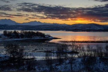 Sonbaharın sonlarında kısa bir günün gündoğumu sırasında kış günü. Karla kaplı topraklar. Renkli gökyüzü. Norveç denizinin suyu ve arka planda dağlar. Sorvika, Norveç.