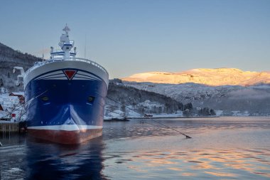 Limanı olan büyük bir tekne. Arkasında güneş ışığı olan karlı dağlar. Mavi gökyüzü. Su manzarayı yansıtıyor. Foldvik, Norveç.