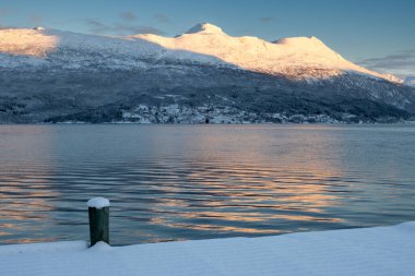 Dağlar bir fiyortta Norveç denizinin sakin suyuyla kaplı. Dağların zirveleri gün ışığıyla aydınlanır. Karla kaplı topraklar. Mavi gökyüzü. Foldvik, Norveç.