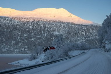 Bir fiyort (Norveç Denizi) kıyısında donmuş su ve arka planda güneş ışığıyla aydınlanan bir nehir. Sahildeki evler. Karla kaplı topraklar. Mavi gökyüzü. Gratangsbotn, Norveç.
