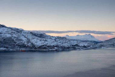Fiyortta sakin su (Norveç denizi). Karlı dağlar. Gün batımında açık gökyüzü ve açık bulutlar renklenmeye başlıyor. Tennevoll, Norveç.