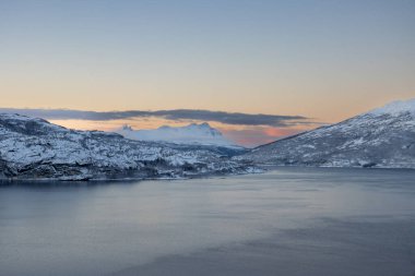 Fiyortta sakin su (Norveç denizi). Karlı dağlar. Gün batımında açık gökyüzü ve açık bulutlar renklenmeye başlıyor. Tennevoll, Norveç.