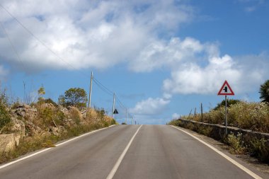 Tepenin yukarısında ağaçlar ve çalılarla kaplı bir yol var. Trafik levhası: soldan bir yol ayrımı. Beyaz bulutlu mavi gökyüzü. Torrefano, Sicilya, İtalya.