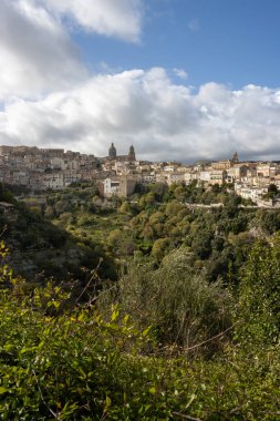 Tepelerde inşa edilmiş ve çevresi yeşilliklerle çevrili eski bir tarihi kasaba. Binaların çatıları var. İlkbaharda yoğun beyaz bulutlu mavi gökyüzü. Ragusa, Sicilya, İtalya.