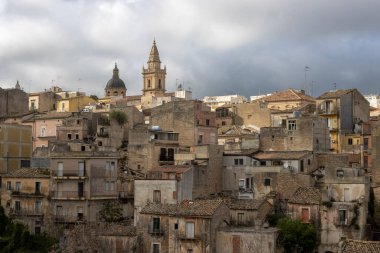 Tepelerde inşa edilmiş ve çevresi yeşilliklerle çevrili eski bir tarihi kasaba. Binaların çatıları var. İlkbaharda yoğun beyaz bulutlu mavi gökyüzü. Ragusa, Sicilya, İtalya.