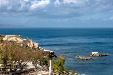 Blue calm water of the Mediterranean sea. Promenade on the coast and several old houses. Mountain on the horizon. Blue sky with white clouds in the spring. Portopalo, Sicily, Italy. clipart