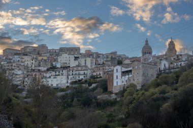 Tepelerde inşa edilmiş ve çevresi yeşilliklerle çevrili eski bir tarihi kasaba. Binaların çatıları var. İlkbaharda yoğun beyaz bulutlu mavi gökyüzü. Ragusa, Sicilya, İtalya.