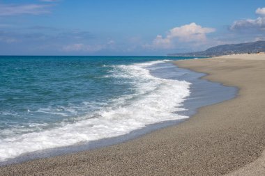 Baharda hiç insan olmayan kumsal. Thyrrenian (Akdeniz) denizinin zümrüt rengi ve ışık dalgaları. Sırtüstü uzanan dağ. Beyaz bulutlu mavi gökyüzü. Spadafora, Sicilya, İtalya.