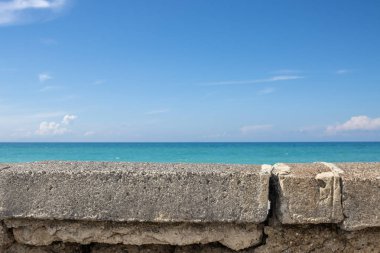 Old weathered concrete fence. Turquoise color of the calm Thyrrenian (Mediterranean) sea with its horizon. Blue sky with light white clouds in the spring. Spadafora, Sicily, Italy. clipart