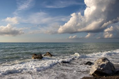 Sahildeki kayalar gün batımının erken ışıklarıyla aydınlandı. Doğanın yumuşak renkleri. Küçük Tyrhenian dalgaları (Akdeniz). Beyaz bulutlu mavi gökyüzü. Villafranca, Sicilya, İtalya.
