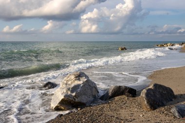Gün batımının erken ışıklarıyla aydınlanan kayalarla dolu kumsal. Doğanın yumuşak renkleri. Küçük Tyrhenian dalgaları (Akdeniz). Beyaz bulutlu mavi gökyüzü. Villafranca, Sicilya, İtalya.