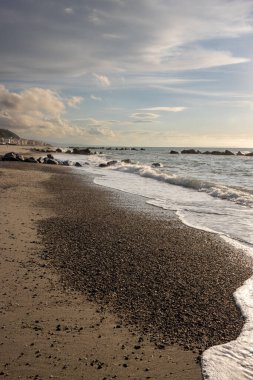 Kumsal, çakıl ve kayalar sahilde, gün batımının erken ışıklarıyla aydınlanmış. Küçük Tyrhenian dalgaları (Akdeniz). Beyaz bulutlu mavi gökyüzü. Villafranca, Sicilya, İtalya.