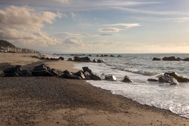 Gün batımının erken ışıklarıyla aydınlanan kayalarla dolu kumsal. Doğanın yumuşak renkleri. Küçük Tyrhenian dalgaları (Akdeniz). Beyaz bulutlu mavi gökyüzü. Villafranca, Sicilya, İtalya.