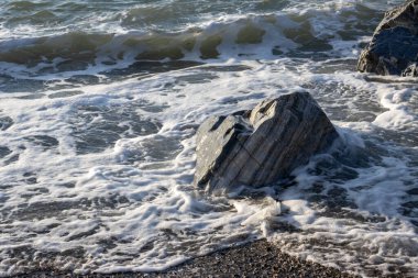 Kıyı şeridindeki çakıllar gün batımının erken ışıklarıyla aydınlanan kayalarla aydınlanıyor. Doğanın yumuşak renkleri. Küçük Tyrhenian dalgaları (Akdeniz). Villafranca, Sicilya, İtalya.