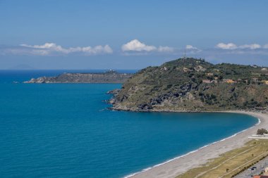 Akdeniz 'in sakin sularının parlak koyu turkuaz rengi (Akdeniz). Sahilde sahil, ardından kayalar. Beyaz bulutlu mavi gökyüzü. Milazzo, Sicilya, İtalya.